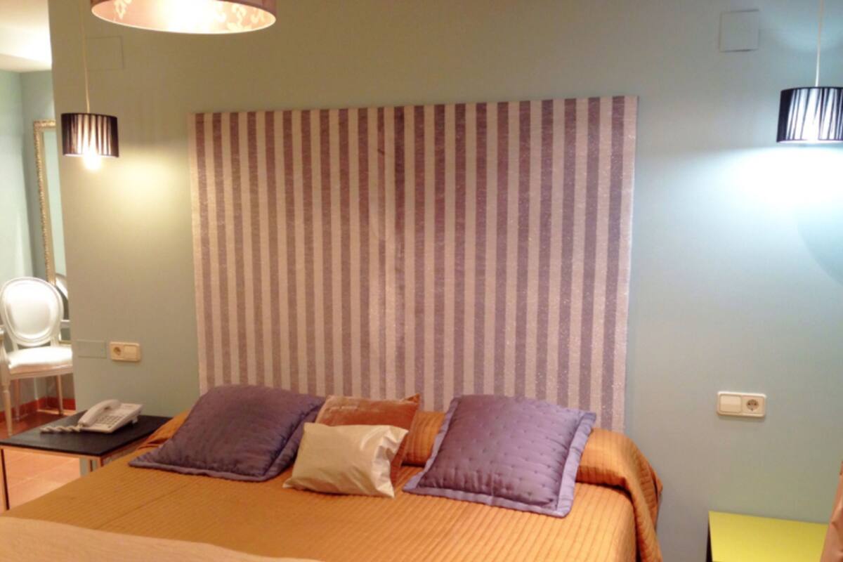 A bedroom with a striped headboard, a bed with two pillows, a bedside table with a lamp, and a chair in the corner.