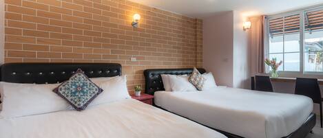 A hotel room with two beds, a brick wall, a desk, and a window with blinds.
