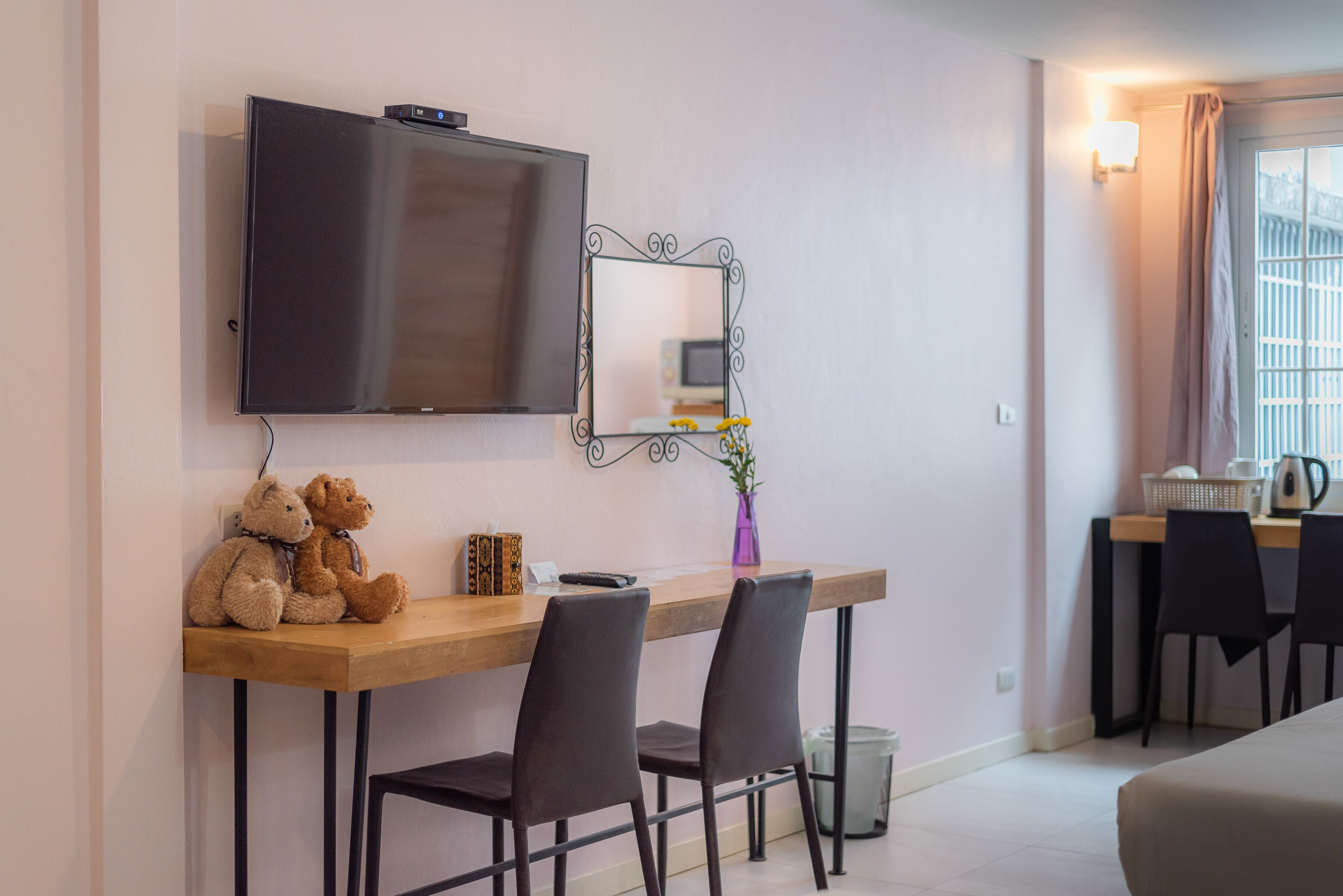 A hotel room with a wooden desk, two chairs, a television mounted above the desk, a small mirror, a vase with flowers, and a window with curtains.