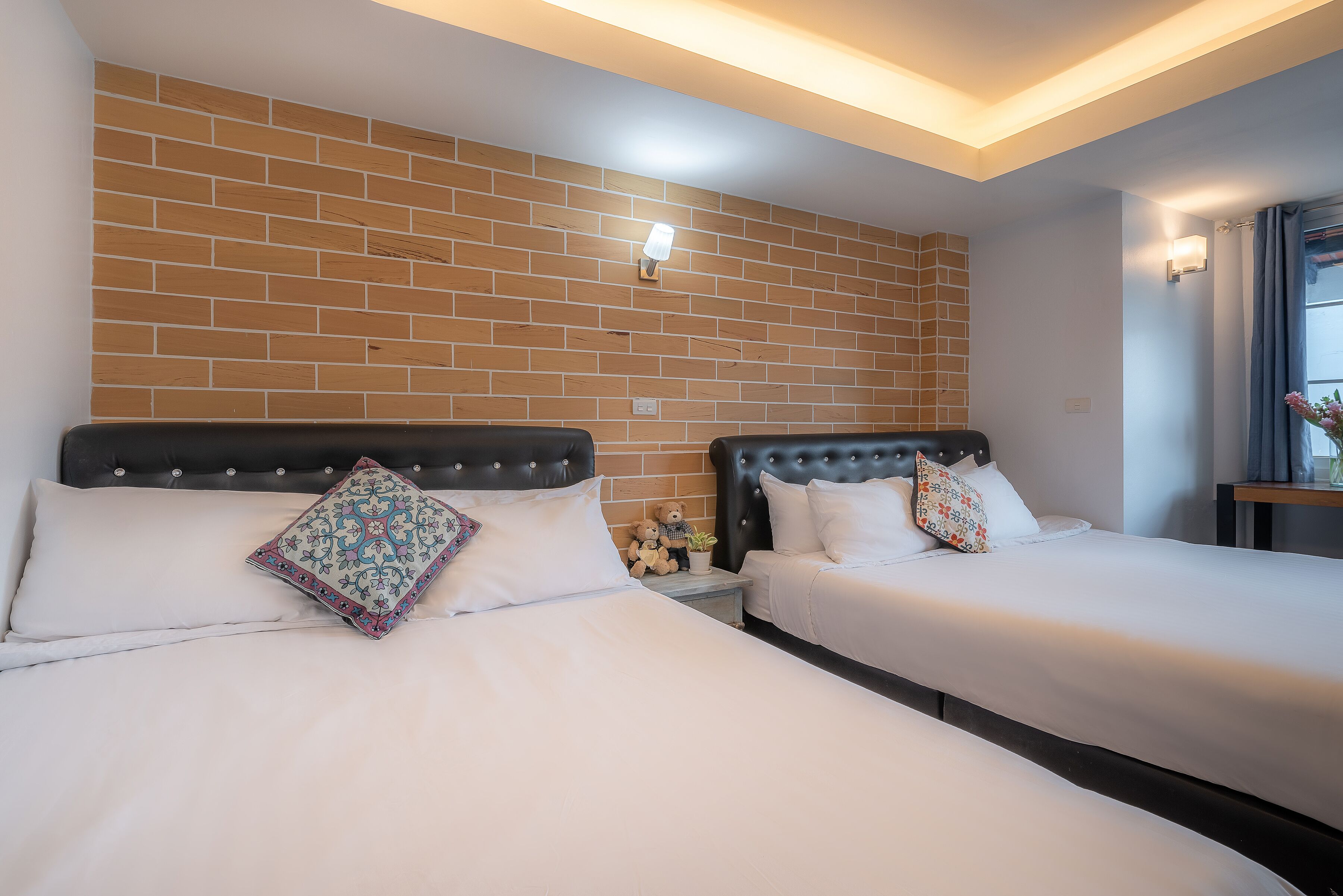 A hotel room with two beds, a brick wall, and a small table with a teddy bear and a plant.