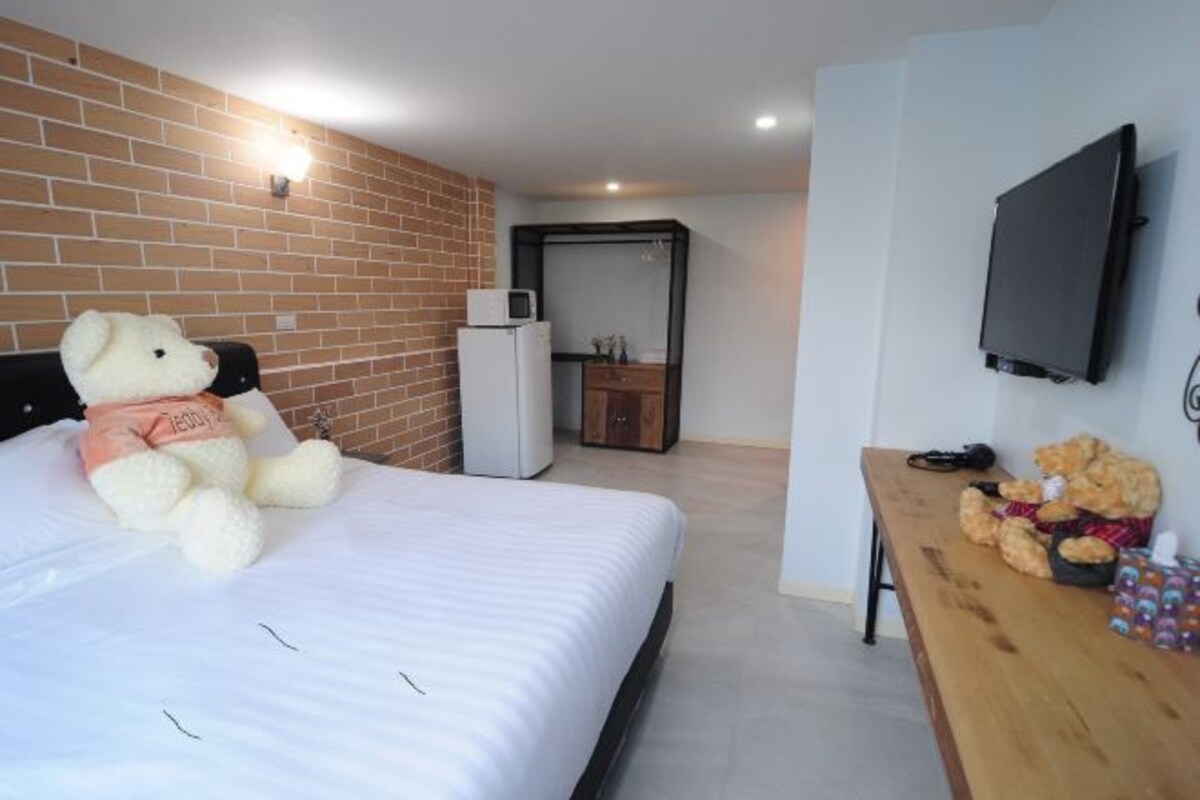 A hotel room with a large bed, a teddy bear, a small refrigerator, a wooden bench, and a flat-screen TV.
