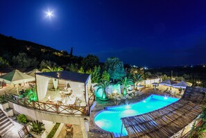 Vista nocturna de un resort con piscina, mobiliario exterior y una zona cubierta.