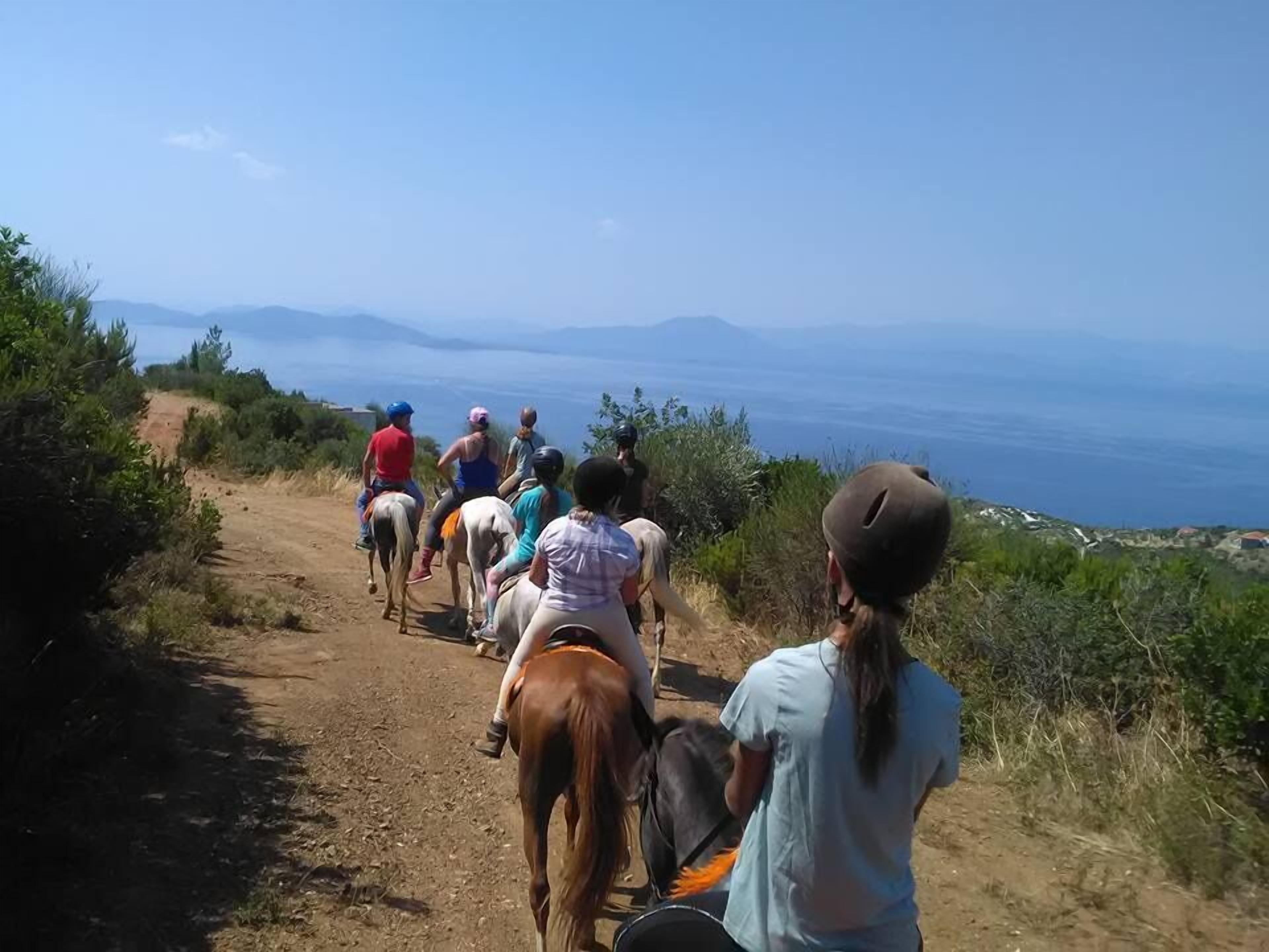 A group of people horseback riding on a dirt path with a view of the sea and distant mountains.
