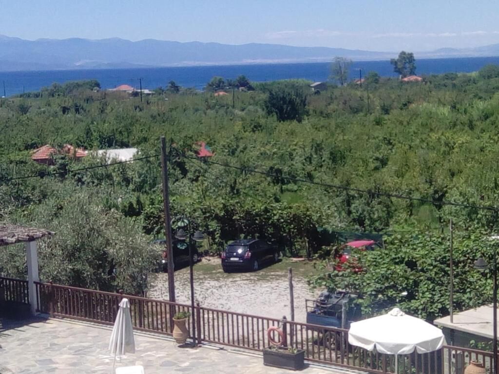 On voit un quartier résidentiel avec une terrasse dallée, une voiture garée près de la maison, et une vue lointaine sur la mer et les montagnes.
