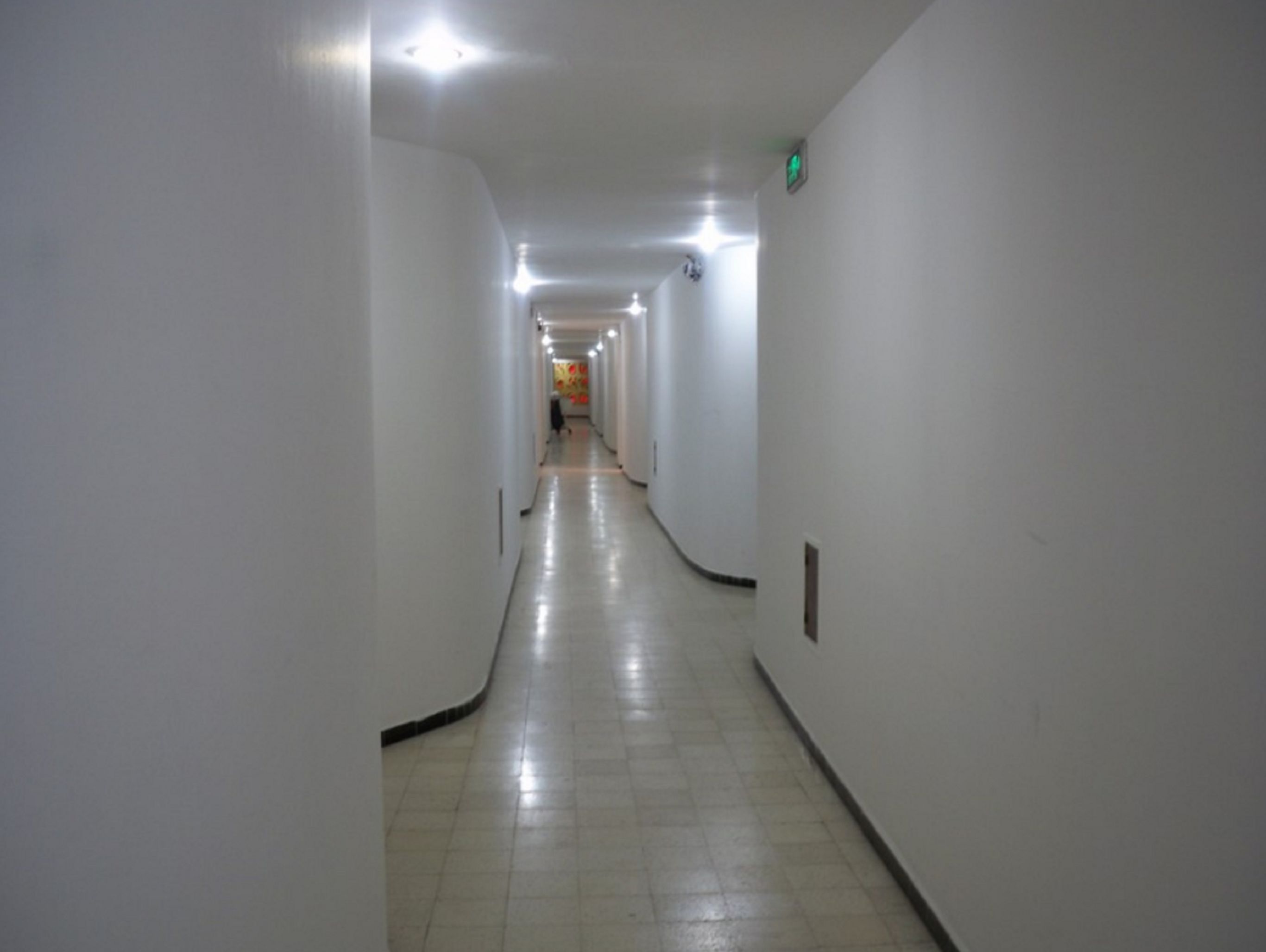A long, narrow hallway with white walls and a tiled floor, leading to a brightly lit room at the end.