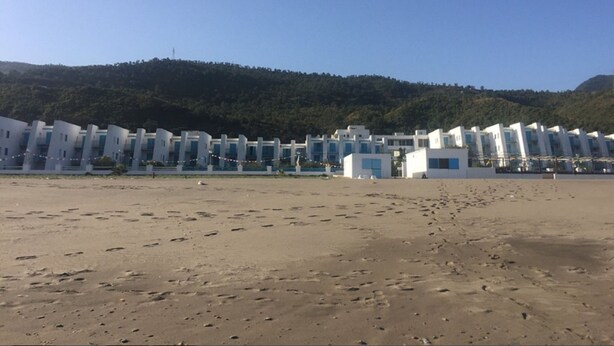 Et hotel ved stranden med en række hvide bygninger, sandstrand i forgrunden og en skovklædt bakke i baggrunden.