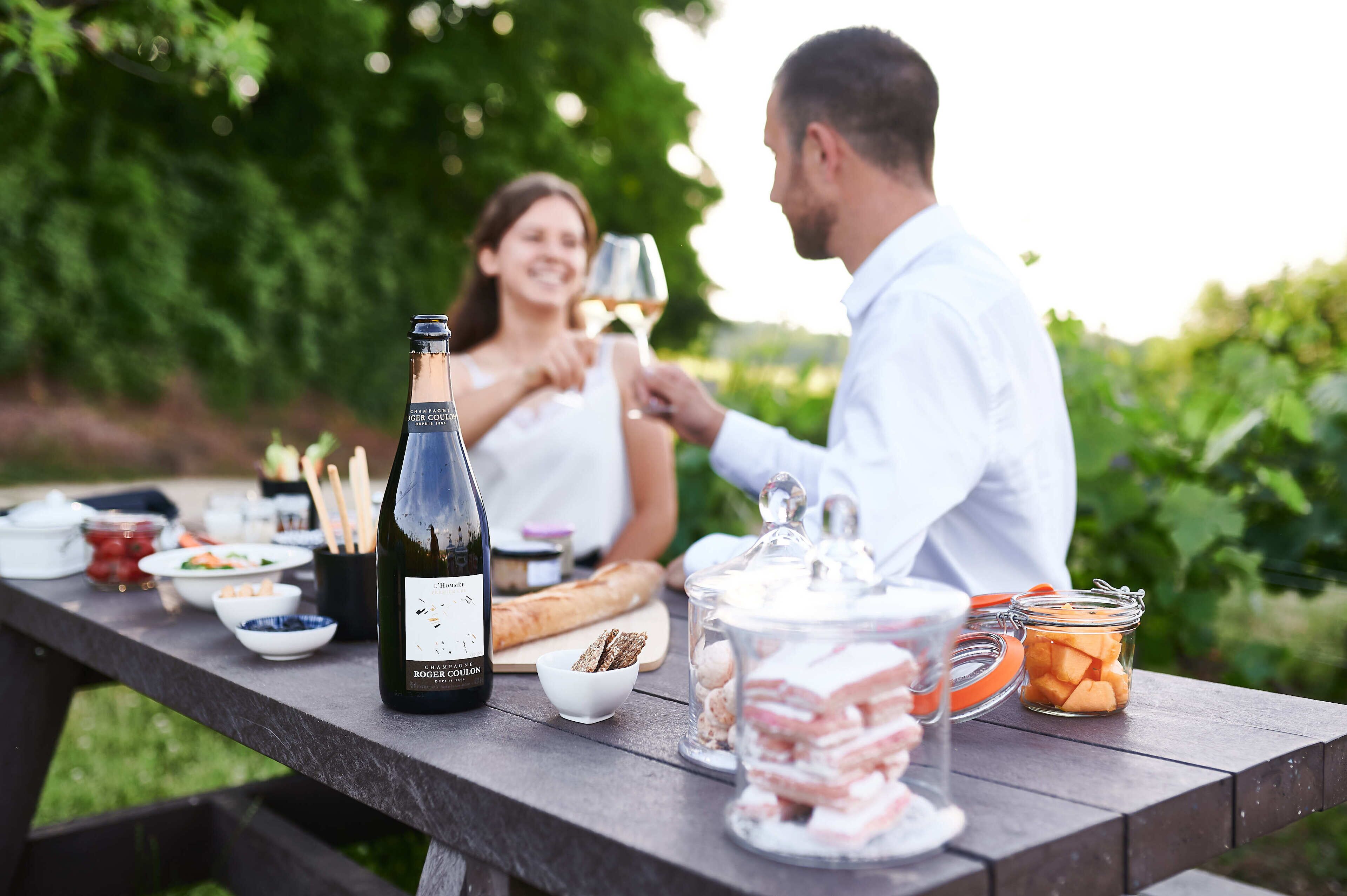A picnic table with food, a bottle of wine, and a glass.