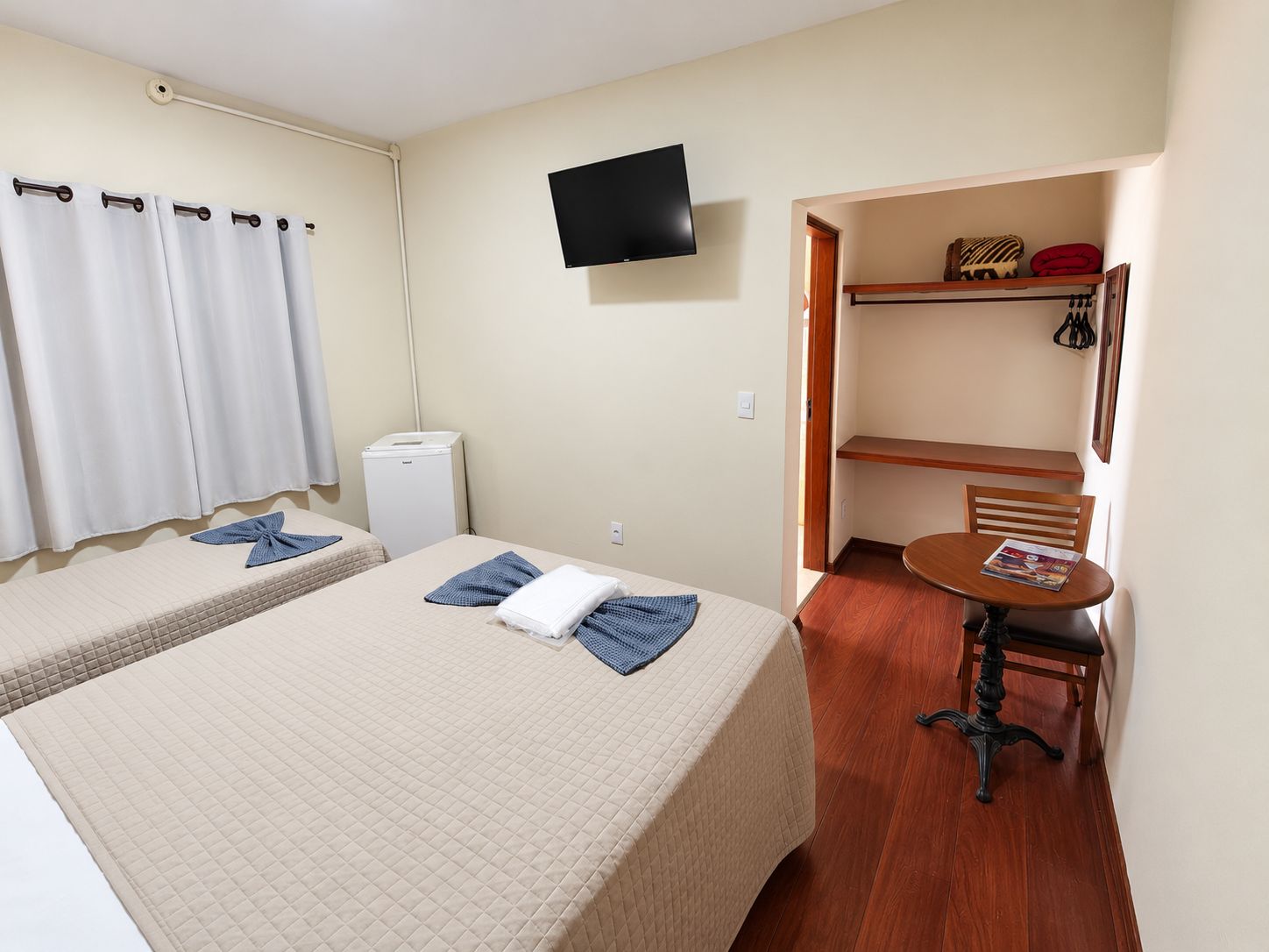 A hotel room with two beds, a television, a small table with a chair, and a shelf with personal items.