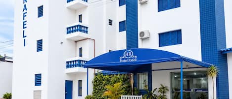 Hotel con fachada blanca y detalles azules, que presenta una entrada cubierta con plantas en macetas y un banco.