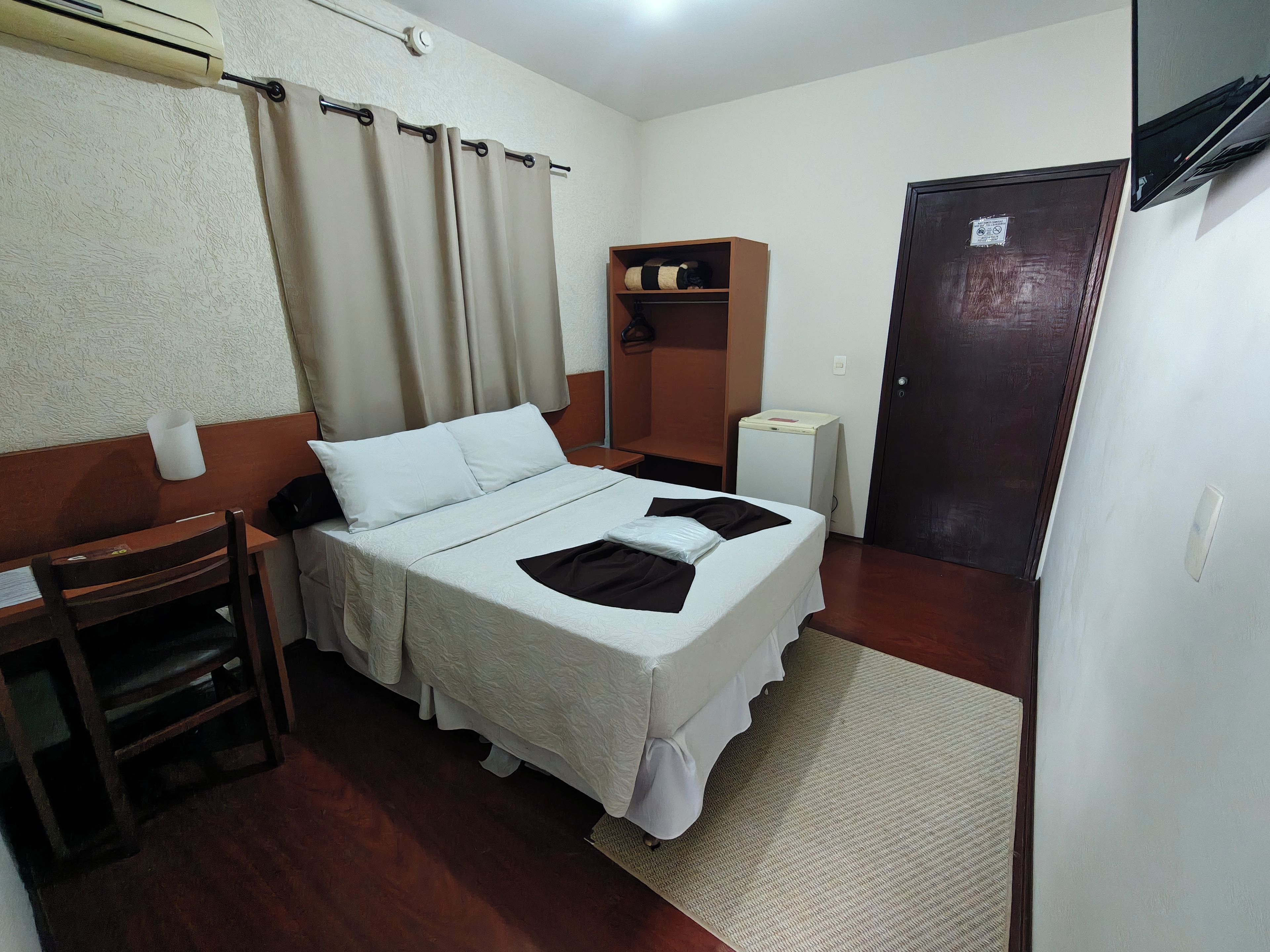 A single bed with white bedding, a wooden nightstand, a small desk with a chair, a wall-mounted air conditioner, a door, and a small cabinet with a shelf.