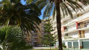 Un hotel a più piani con balconi, un giardino curato con palme e siepi, e un cielo azzurro limpido.