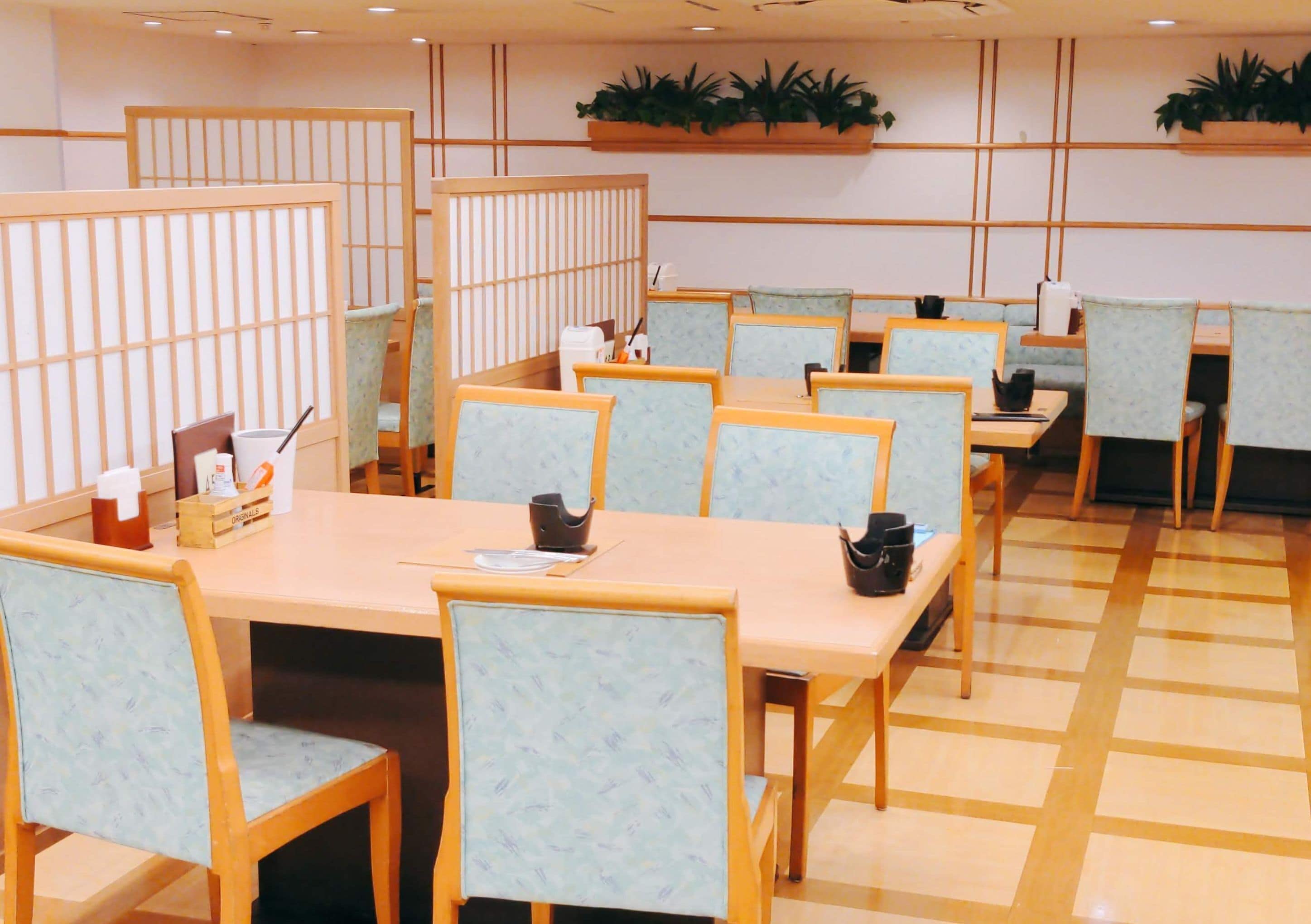 A dining area with wooden tables and chairs, some with cushions. There are plants on the walls and a tiled floor.