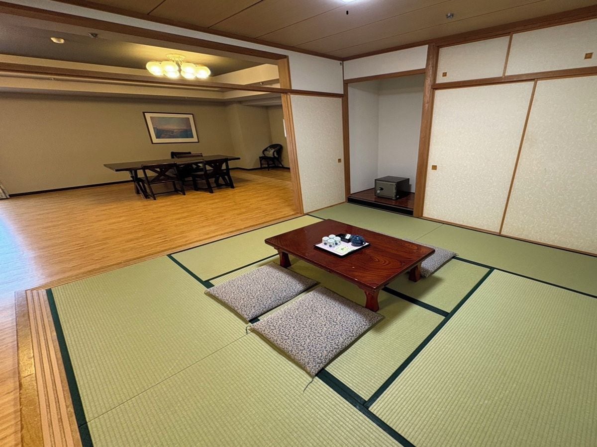 A tatami mat room with a wooden table and cushions, a dining area with chairs and a table in the background, and a sliding door.