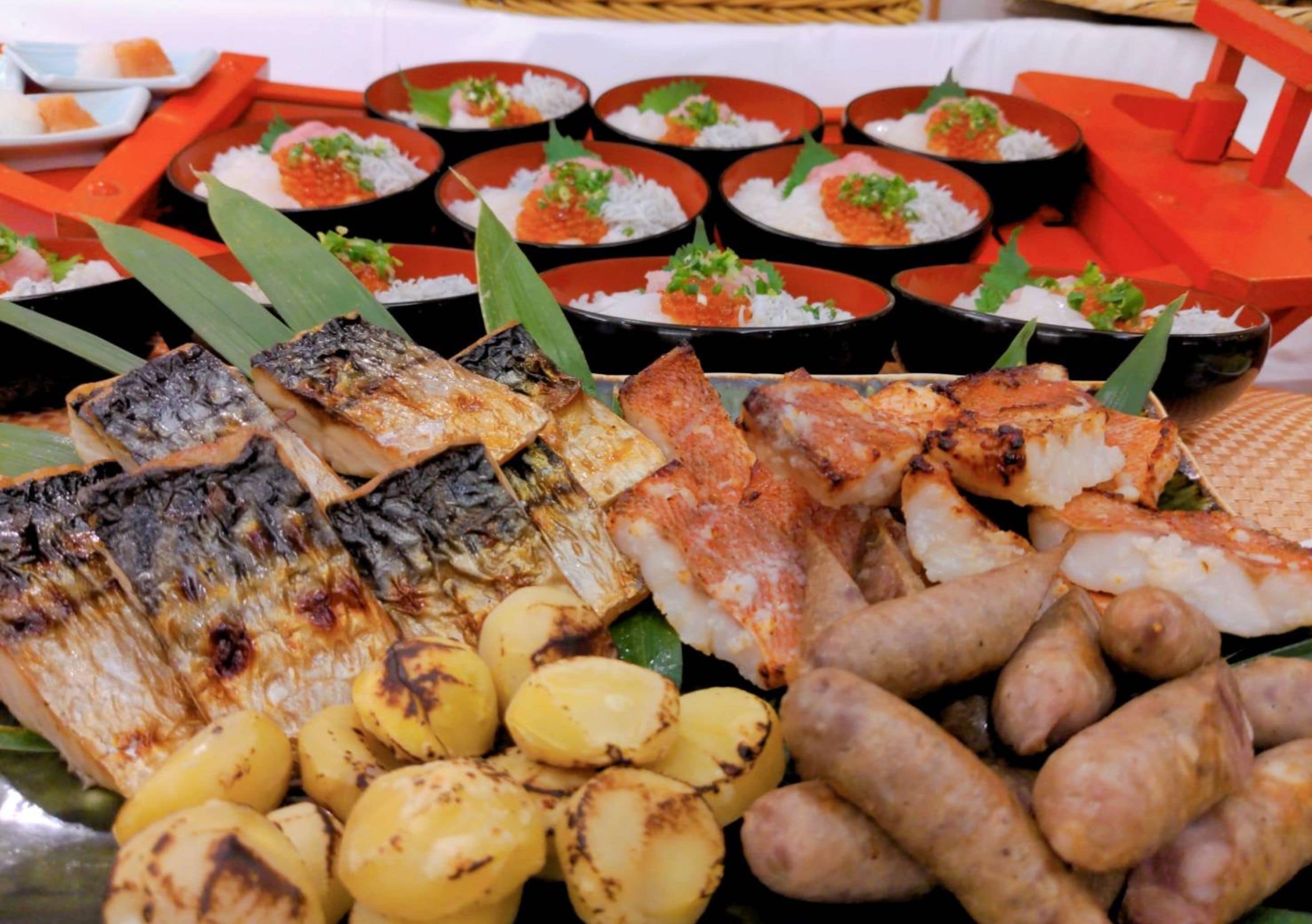 A variety of grilled fish and sausages, with small bowls of rice and a basket in the background.