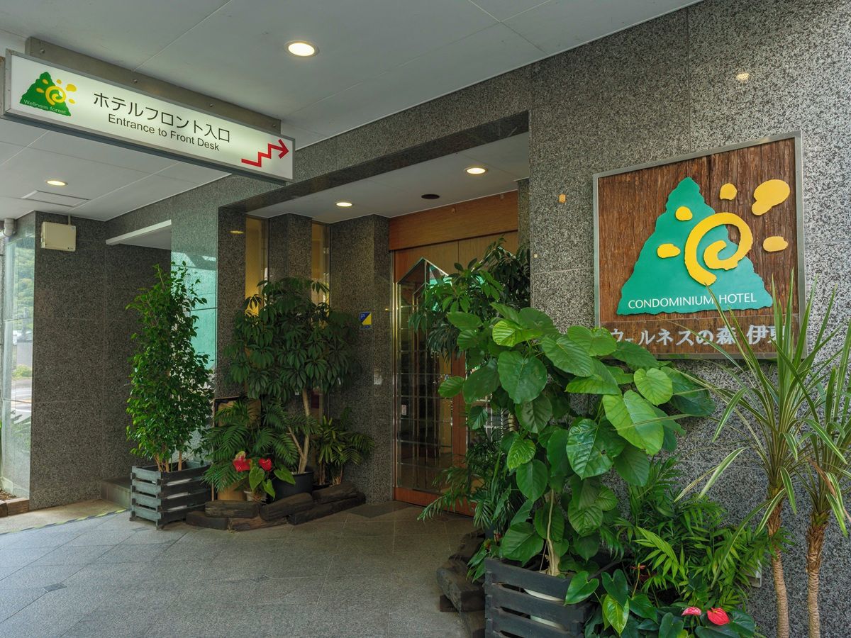 A hotel entrance with a sign indicating the front desk, surrounded by potted plants and a wooden signboard.