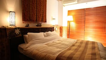 A hotel room with a bed, a nightstand, a lamp, a wall-mounted air conditioner, and a window with blinds.