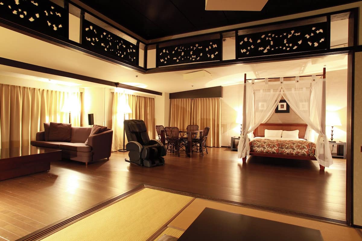 A hotel room with a bed, a massage chair, a sofa, a dining table with chairs, and a large window with curtains.