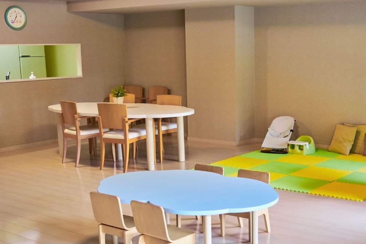 A room with a round dining table, chairs, a massage chair, a play mat, and a small white table with a plant.