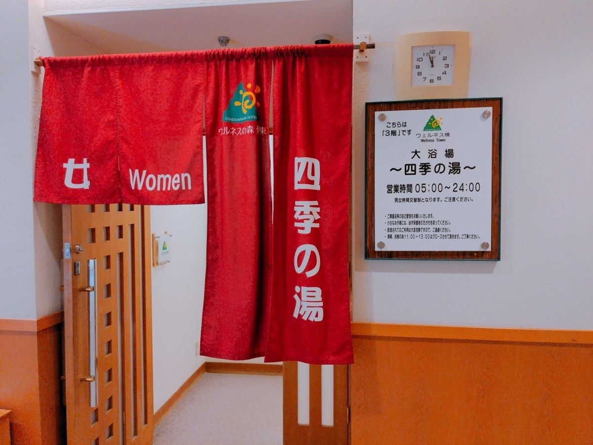 Cortina roja con la frase 'Women' y '4th season bath' escritas, una puerta de madera y un cartel con los horarios de atención.