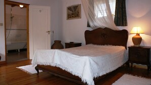 Ein Schlafzimmer mit einem hölzernen Bett, einer Nachttischlampe, einem Nachttisch aus Holz, einem weißen Teppich, einer Holztür und einer Dusche im angrenzenden Raum.