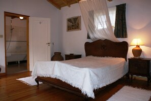 A bedroom with a wooden bed, a bedside lamp, a wooden nightstand, a white rug, a wooden door, and a shower visible in the adjacent room. - Quinta Do Albergue Do Bonjardim (Serta)