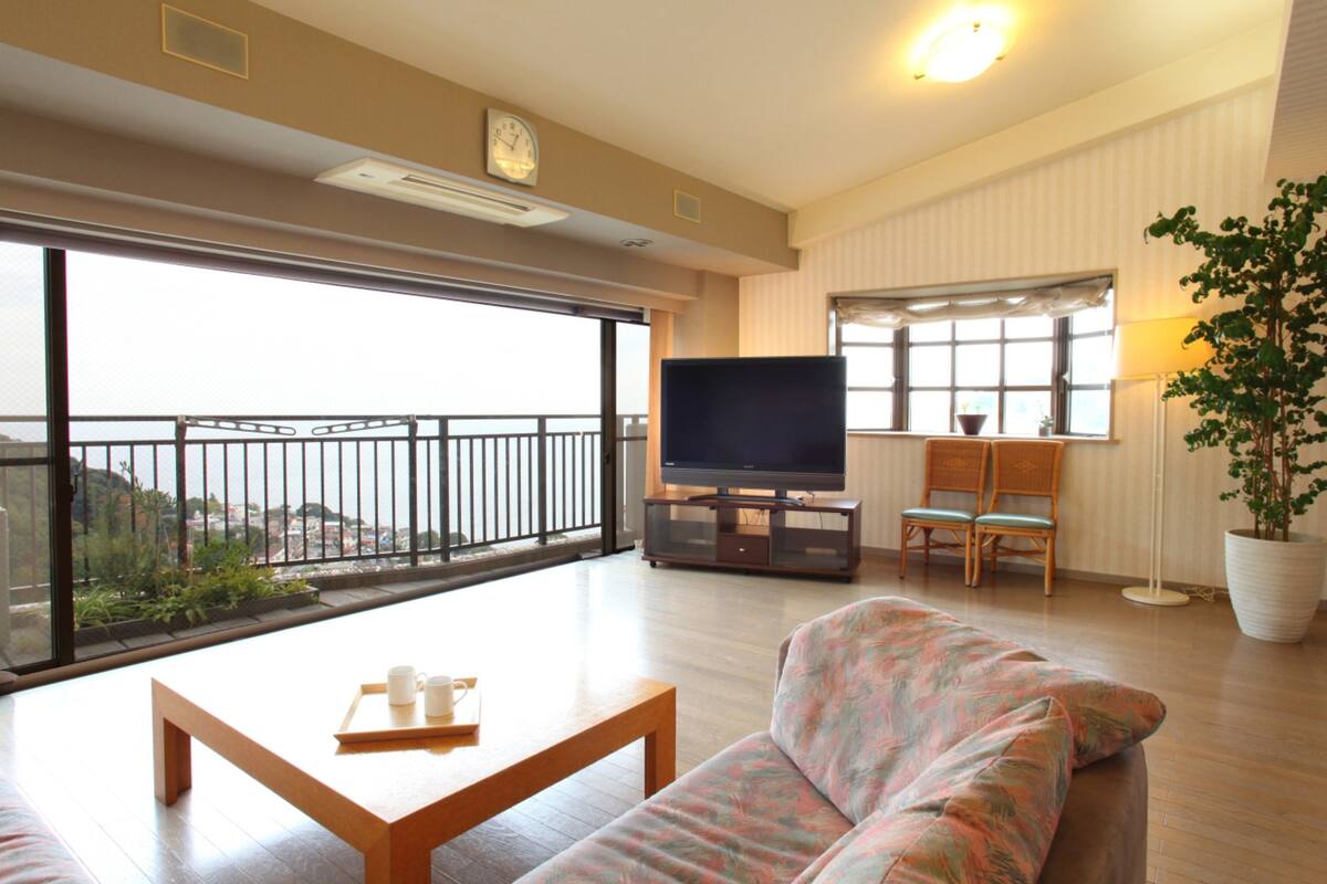 A spacious living room with a large TV, a sofa, a coffee table, and a balcony with a view.