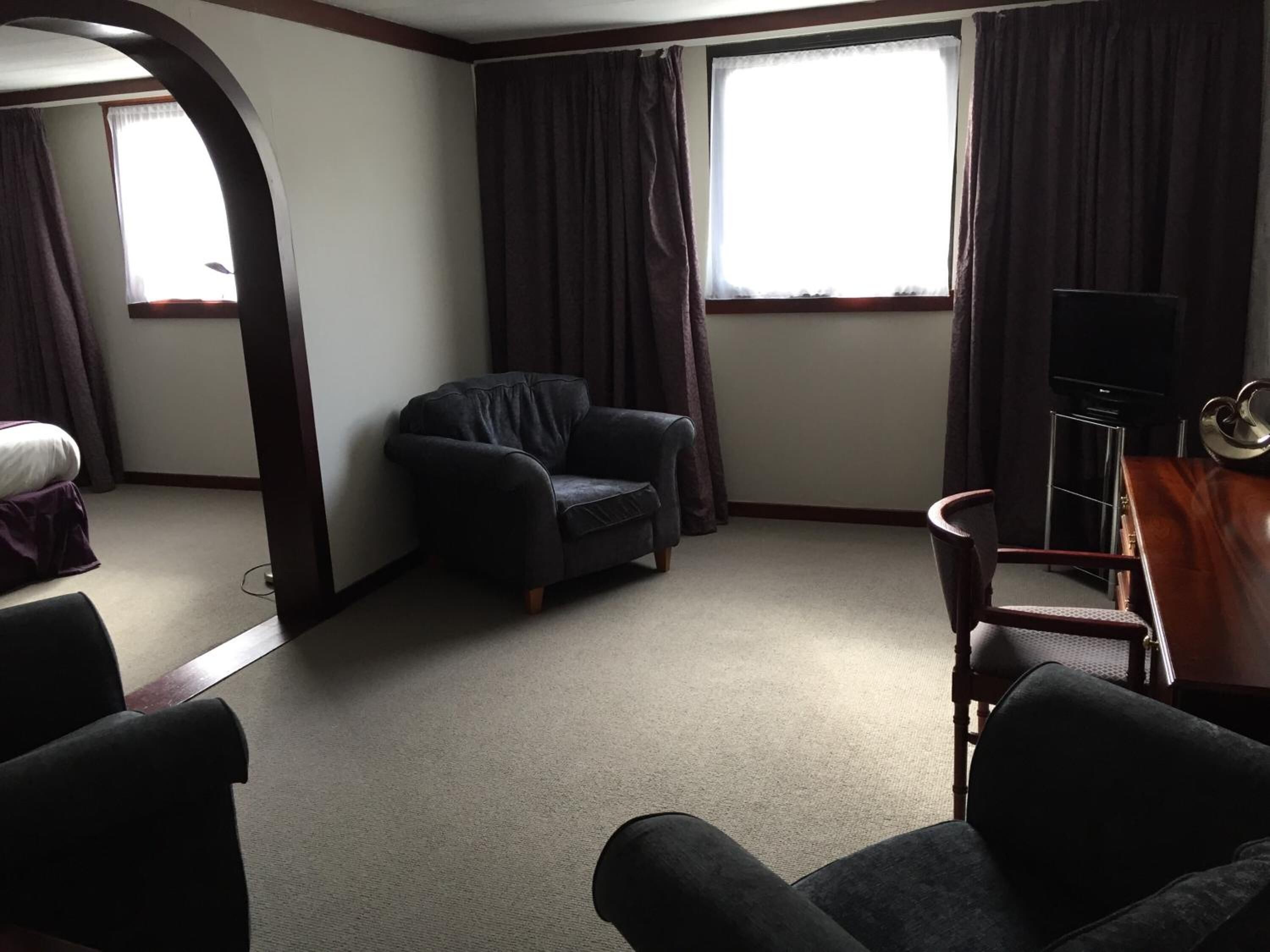 A hotel room with a dark leather armchair, a wooden side table with a television, and a large window with curtains.