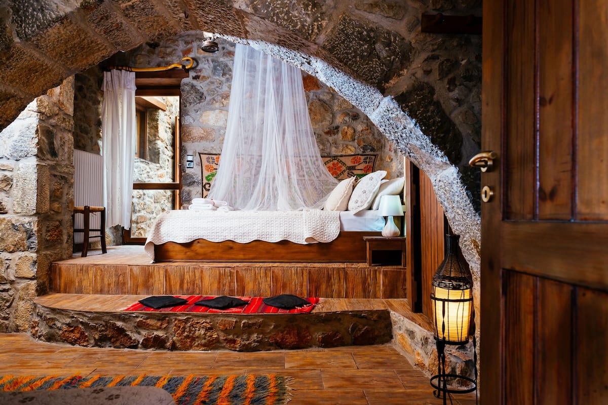 Quarto com paredes de pedra, uma cama, uma mesinha de cabeceira e um candeeiro aceso.