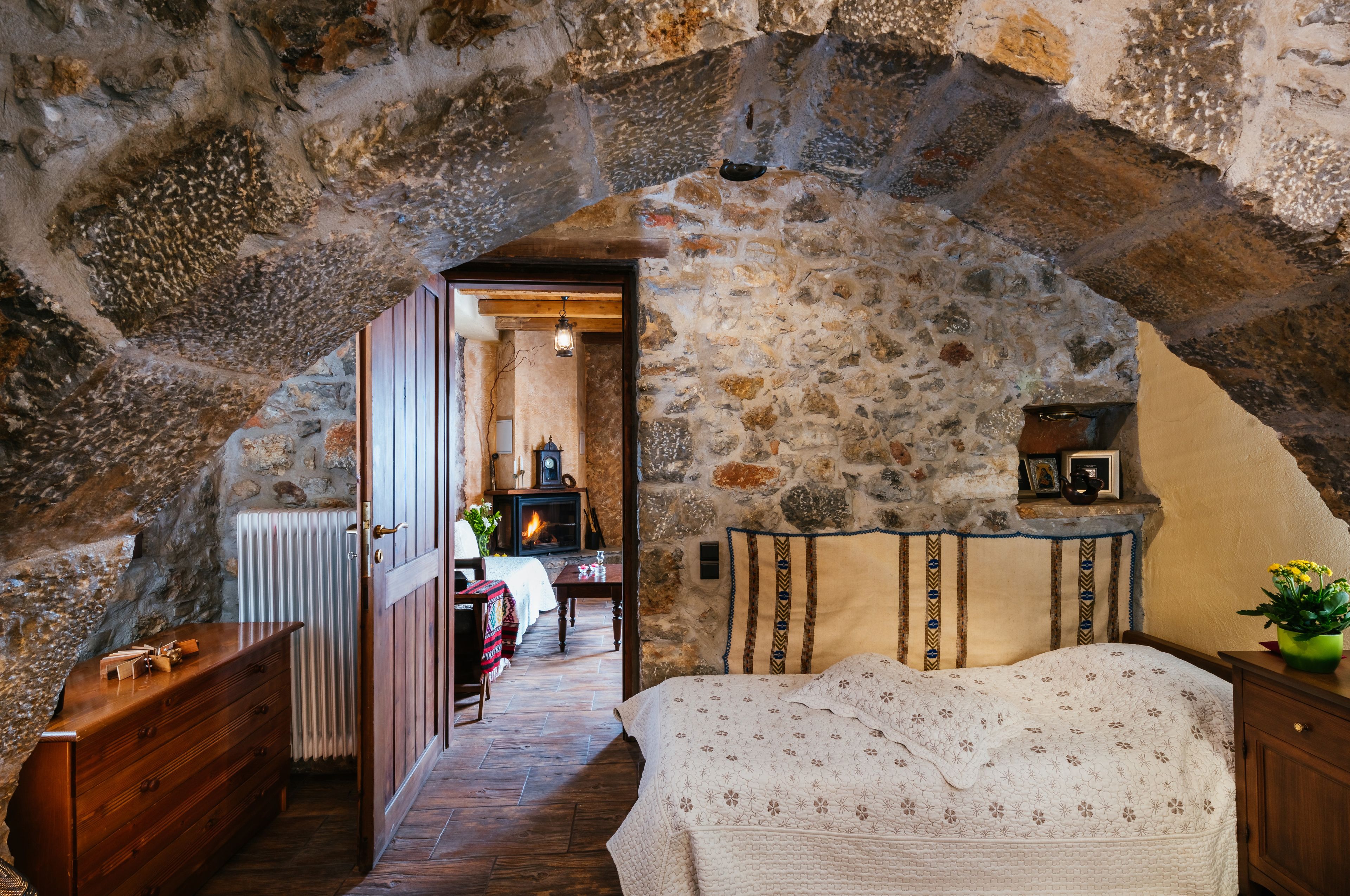 A rustic stone-walled bedroom with a bed, wooden dresser, and a fireplace in the background.
