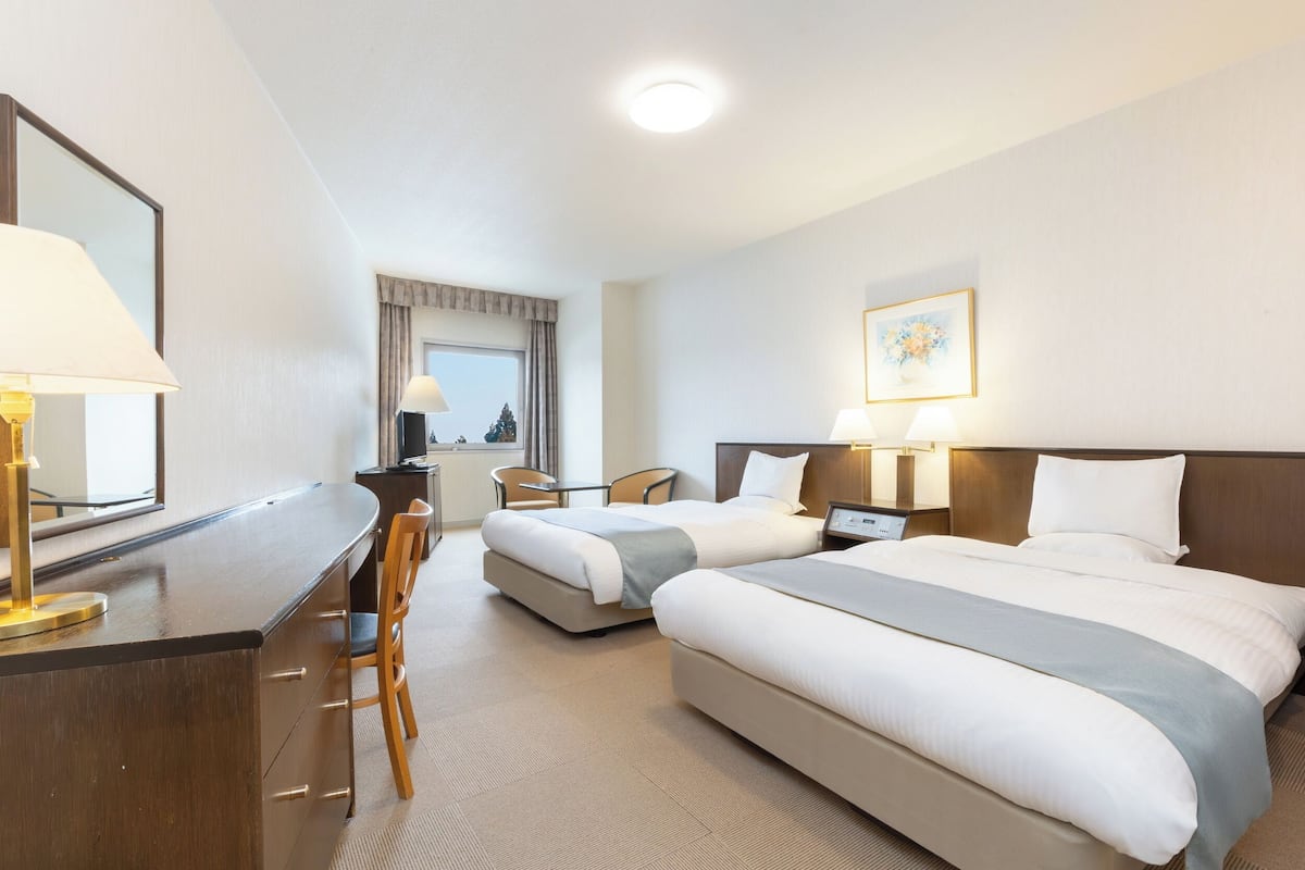 Ett hotellrum med två sängar, ett skrivbord, en stol, ett nattduksbord och ett fönster med gardiner.