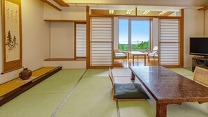 Una habitación japonesa tradicional con suelo de tatami, una mesa baja, un sofá, un televisor y una puerta corredera que da a un balcón con vistas.