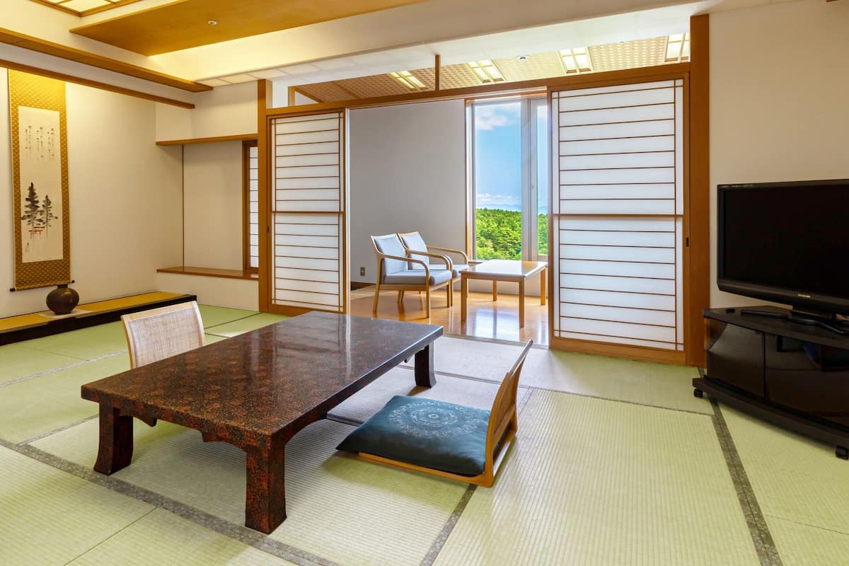 Ett traditionellt japanskt rum med tatamimattor på golvet, ett lågt bord, en soffa, en TV och en skjutdörr som leder ut till en balkong med utsikt.