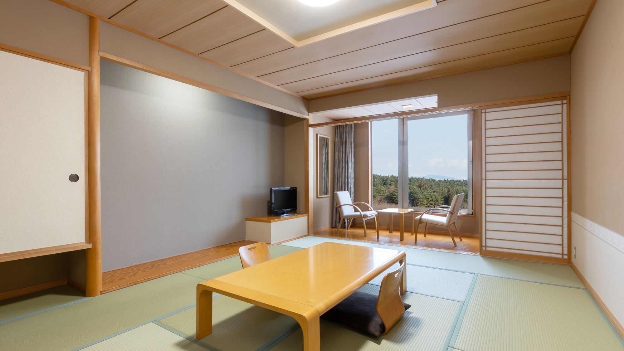 Una habitación con suelo de tatami, una mesa de madera, un televisor y vistas a un bosque a través de amplios ventanales.