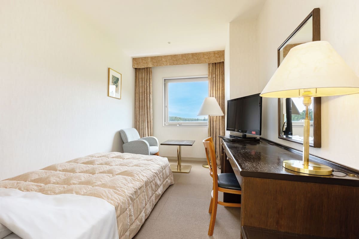 Ett hotellrum med en säng, ett skrivbord med en lampa, en stol, en tv och ett fönster med gardiner.