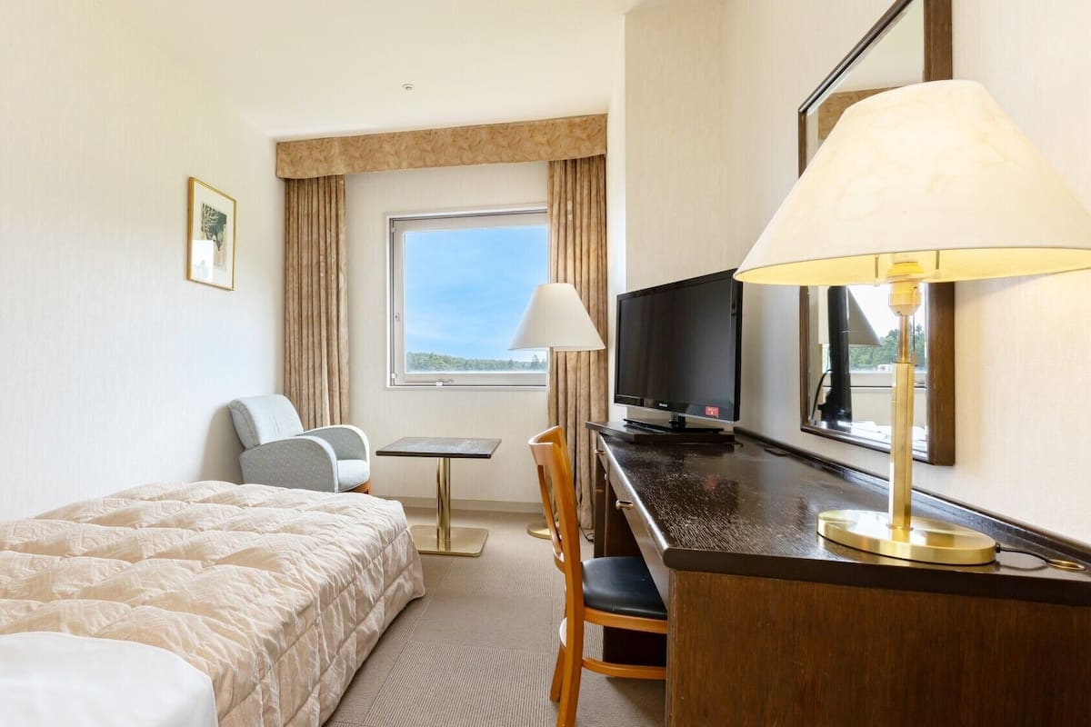 Ett hotellrum med en säng, ett skrivbord, en stol, en tv och ett fönster med gardiner.