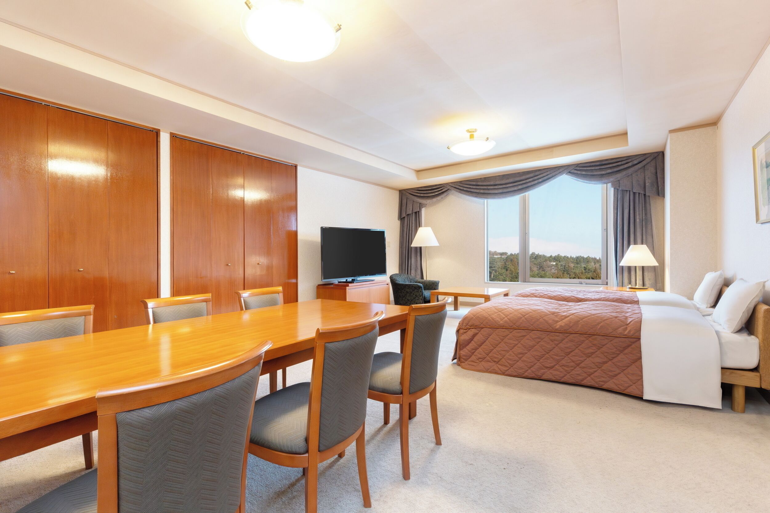 A hotel room with a wooden dining table, four chairs, a bed with a brown quilt, a television, and a large window with curtains.