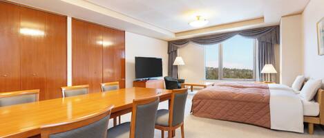 A hotel room with a wooden dining table, four chairs, a bed with a brown quilt, a television, and a large window with curtains.