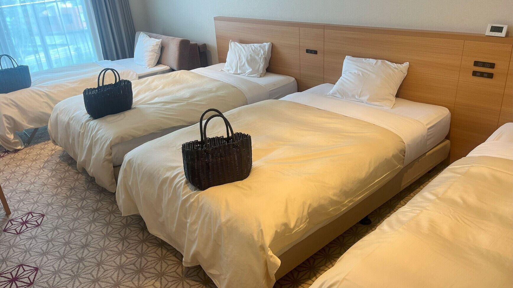 Habitación de hotel con tres camas, cada una con cubrecama y almohadas blancas, y cabeceros de madera. Hay dos bolsas tejidas negras sobre las camas.