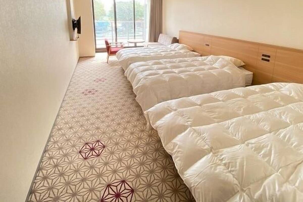 A hotel room with two beds, a patterned floor, a view through a window, and a small table with chairs.