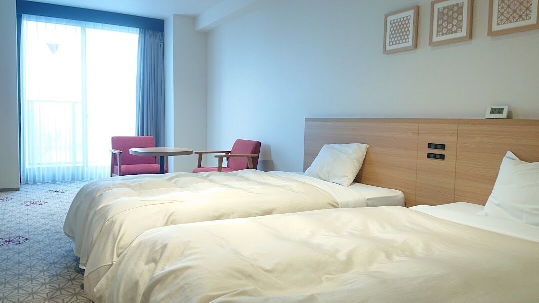 Una habitación de hotel con dos camas, una mesita, una silla y un ventanal grande.
