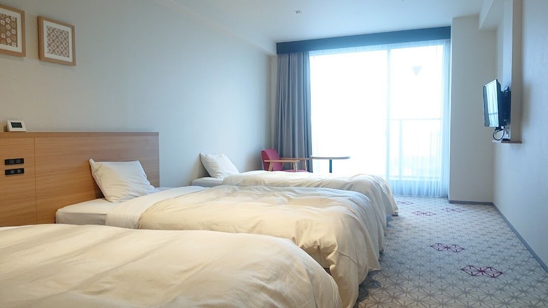 Habitación de hotel con dos camas, una mesita pequeña, una silla, un televisor y un amplio ventanal con cortinas.
