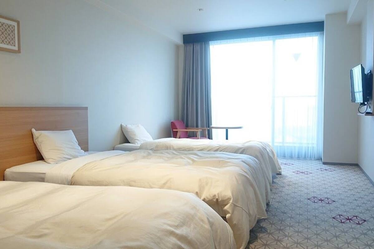 A hotel room with two beds, a small table, a chair, a TV, and a large window with curtains.