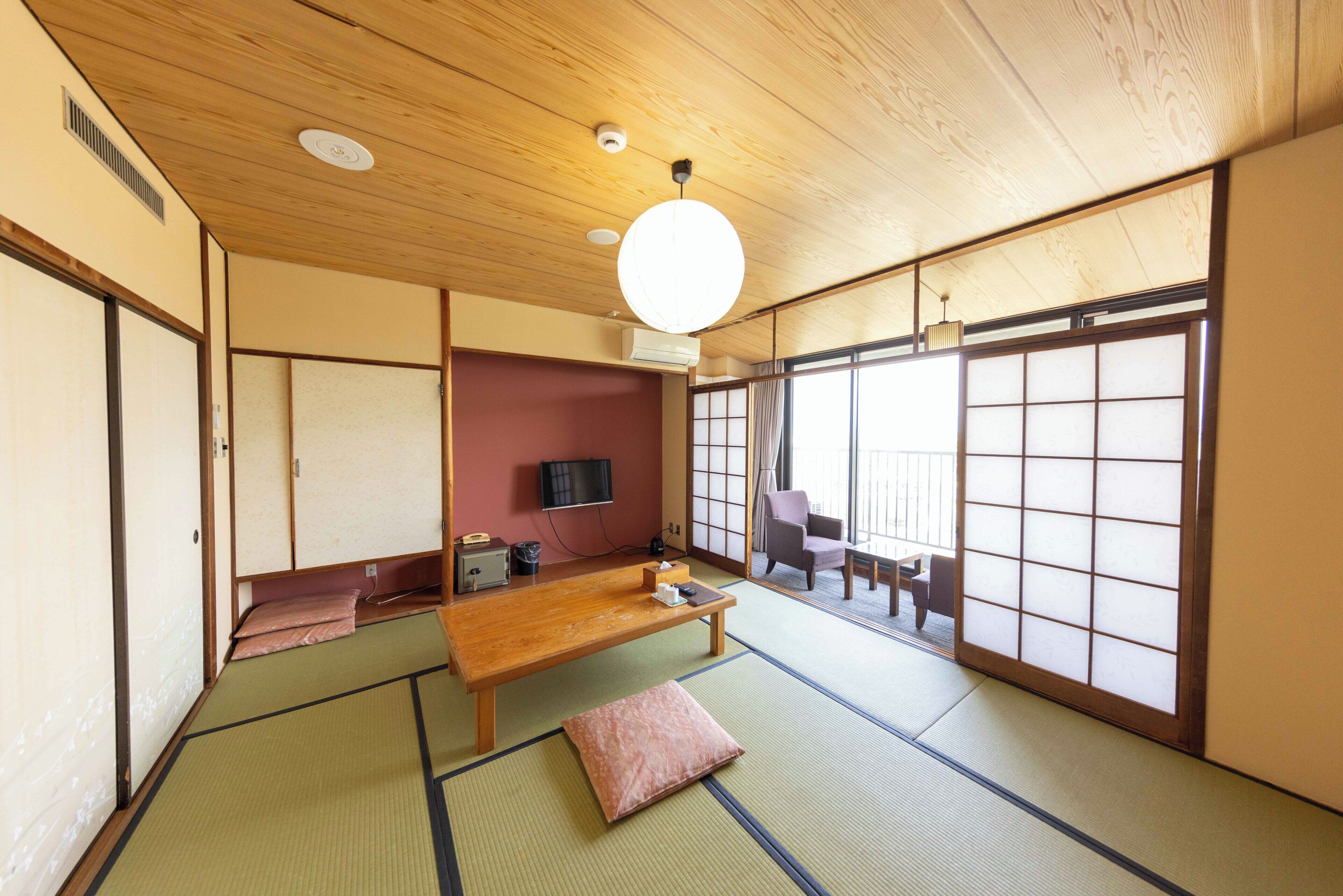 Una habitación japonesa tradicional con suelo de tatami, una mesa de madera baja, un banco acolchado, un televisor y puertas correderas.