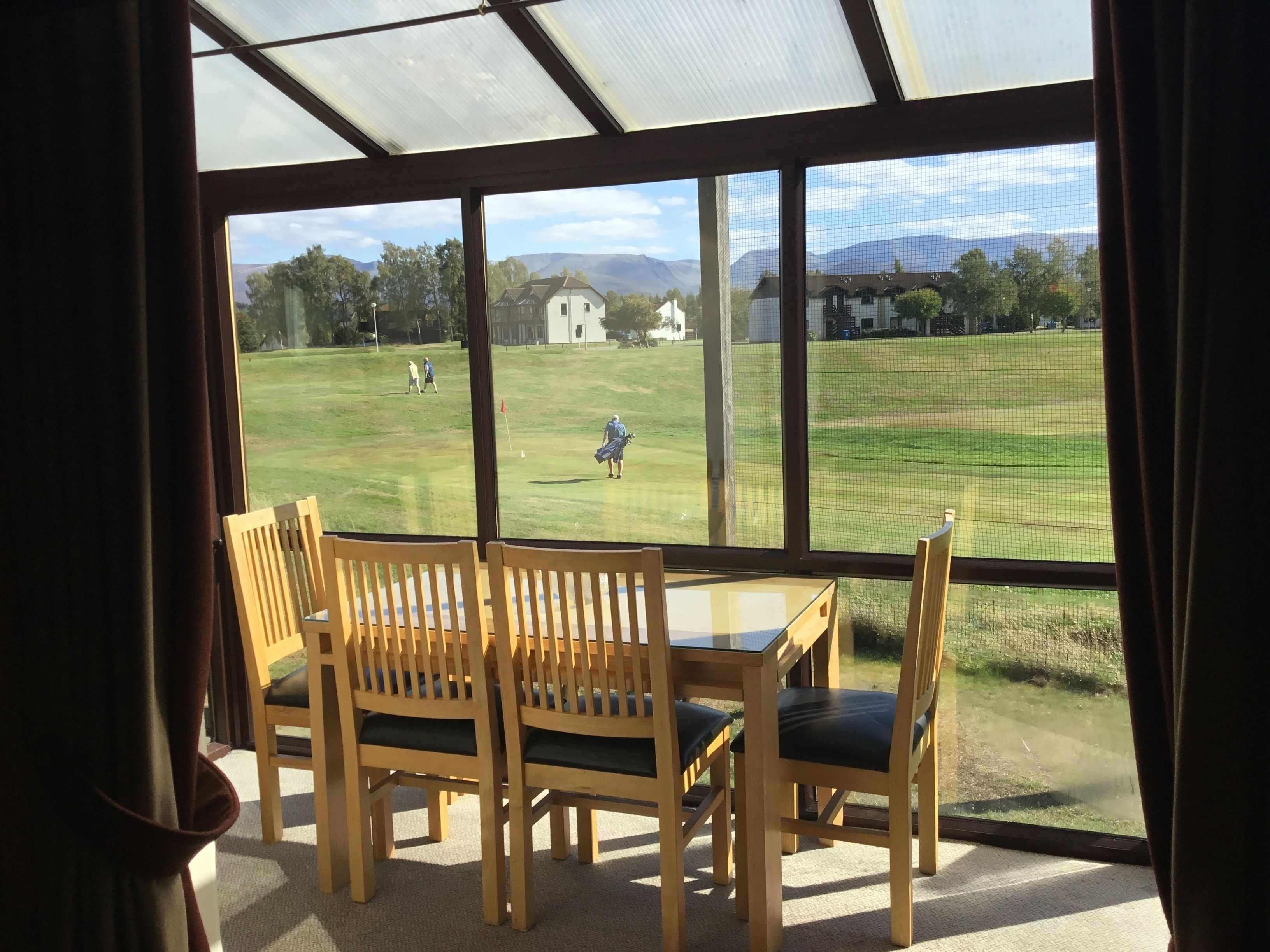 Une pièce dotée d’une grande fenêtre donnant sur un espace verdoyant, avec une maison et des montagnes visibles au loin. À l’intérieur, on trouve des chaises et une table en bois.