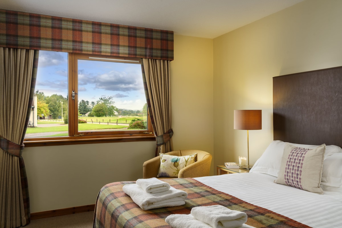 Habitación de hotel con cama, una silla, una lámpara y vistas a un paisaje verde por la ventana.