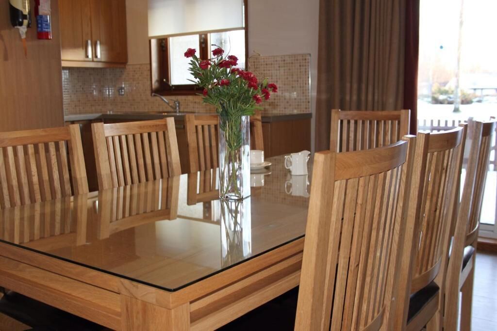 Une table à manger en bois, entourée de six chaises, un vase avec des fleurs rouges et, au fond, une cuisine.