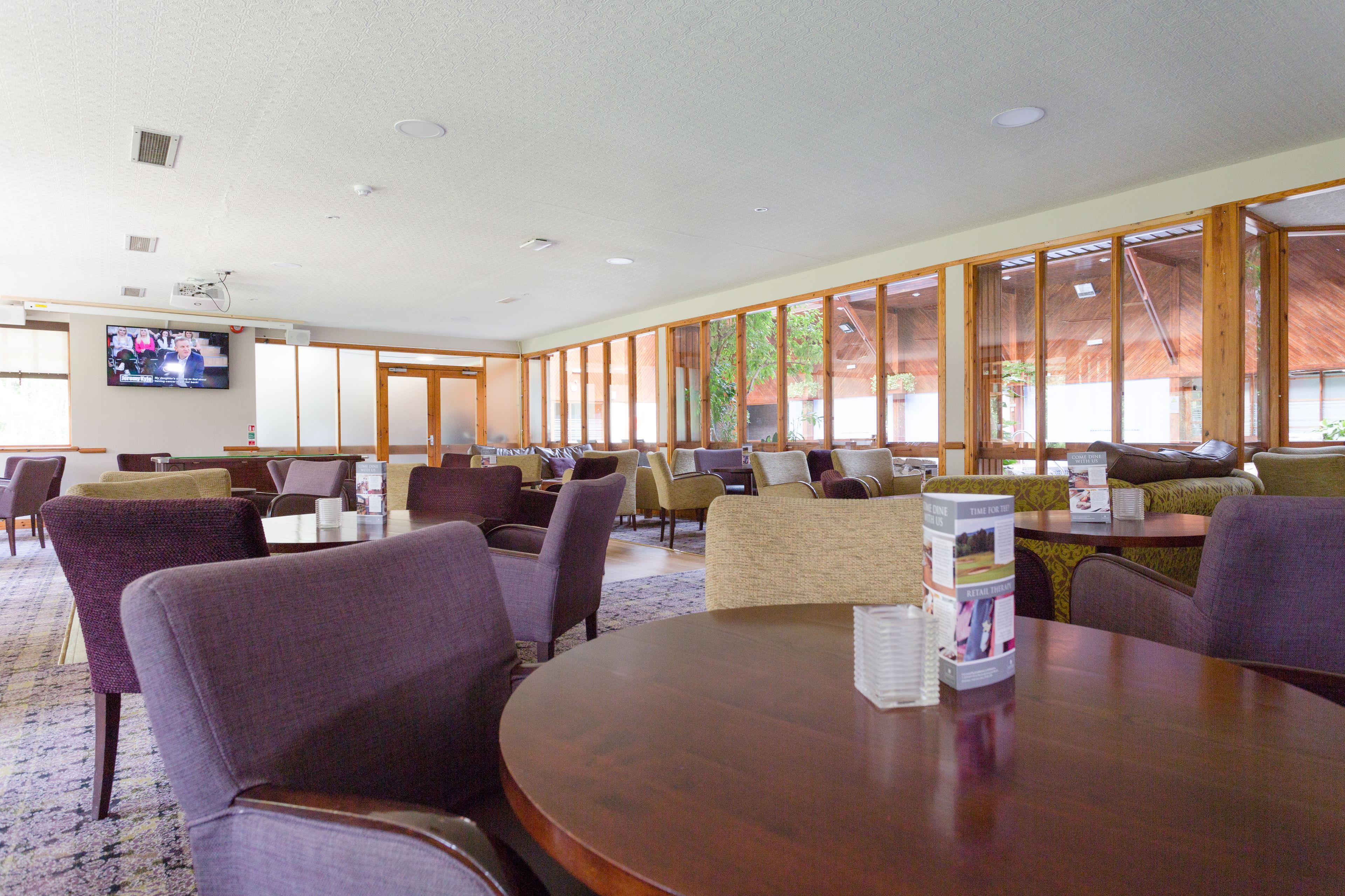 A hotel lobby with a large seating area, a television mounted on the wall, and a view of the outdoors through large windows.