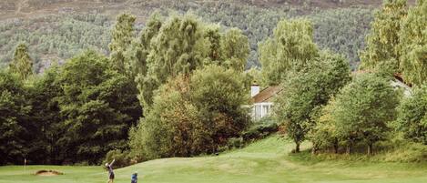 Een golfbaan met een huis op de achtergrond, omringd door bomen en heuvels.