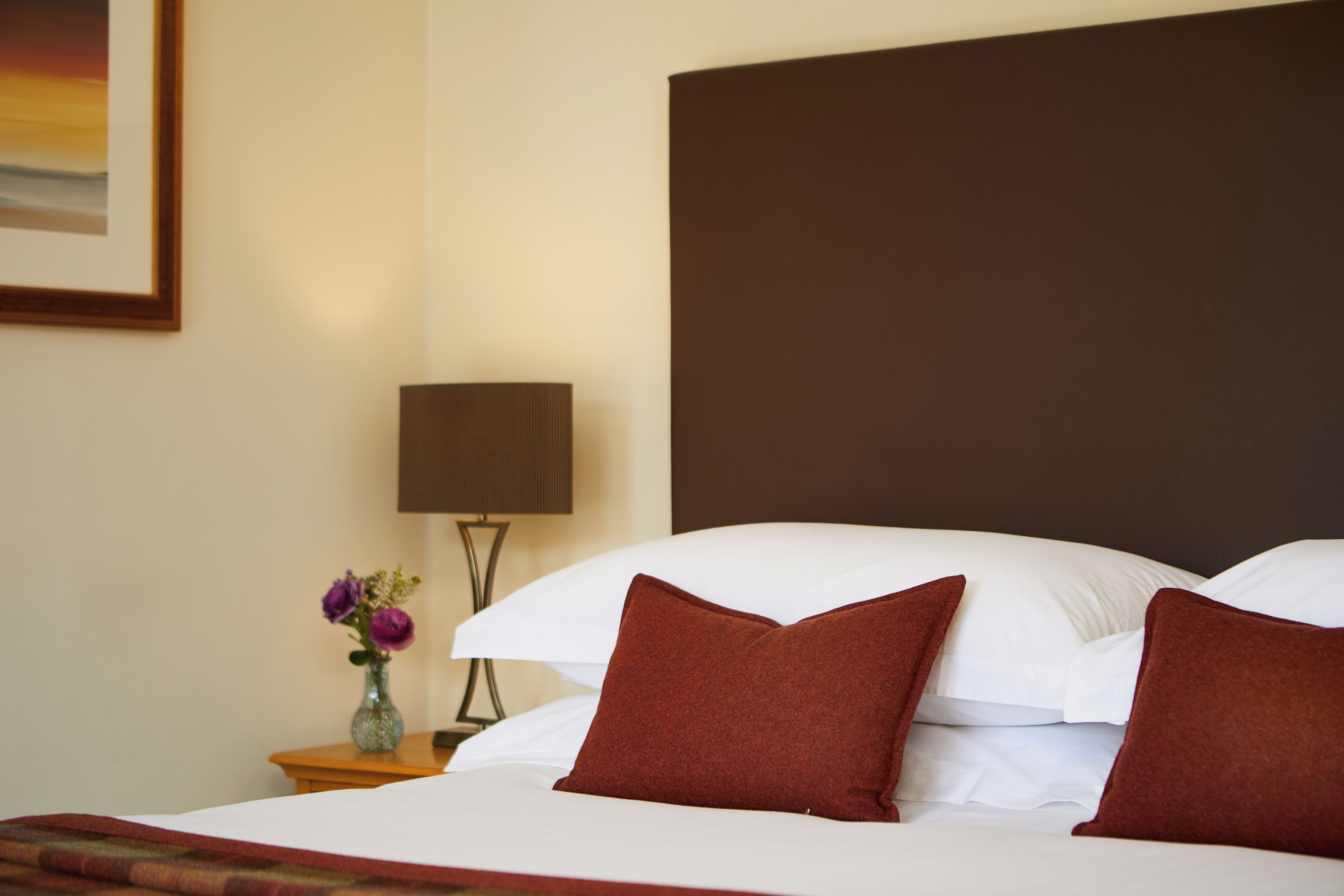 A hotel room with a bed, pillows, a vase with flowers, a lamp, and a painting on the wall.
