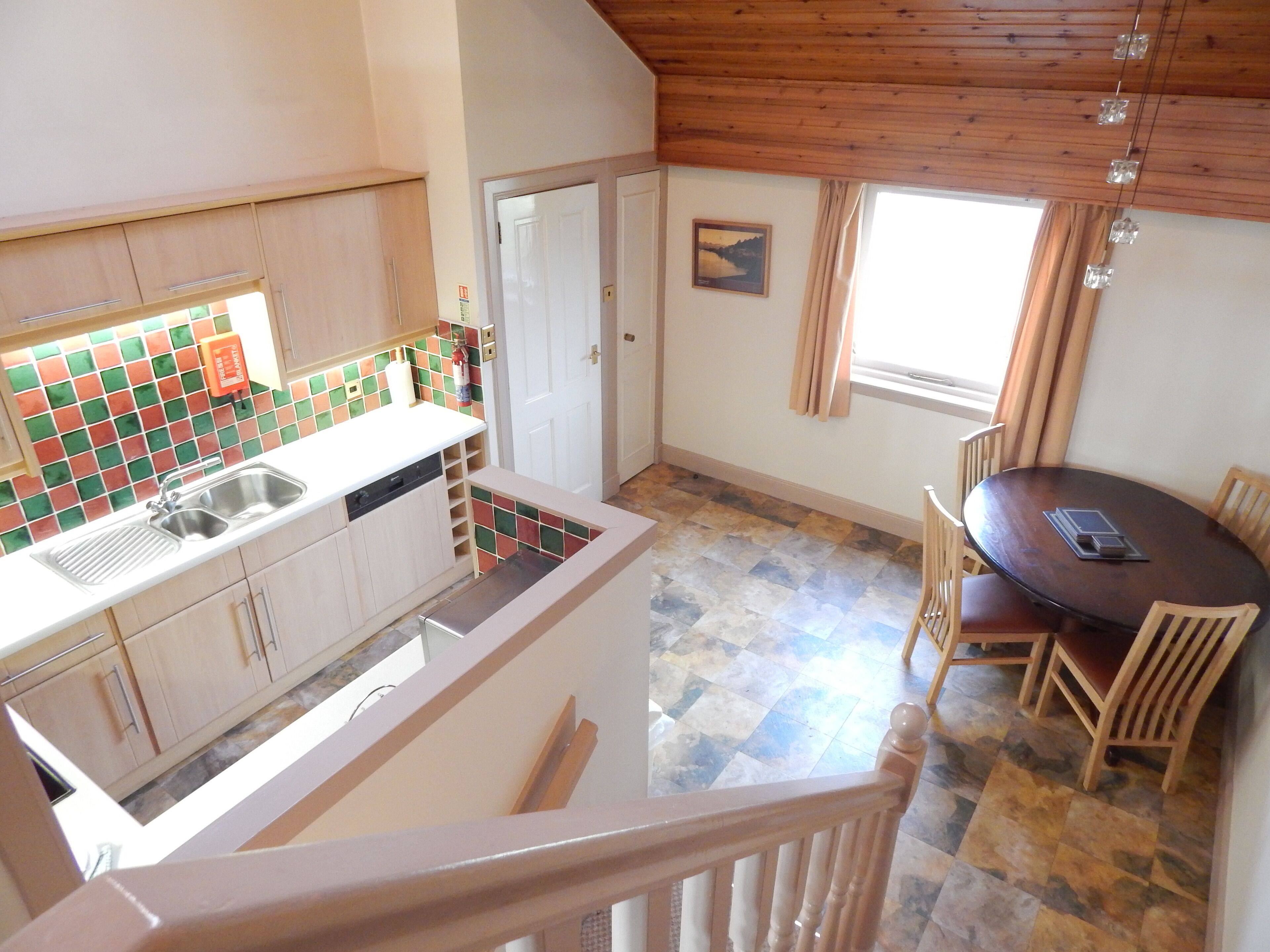 A kitchen with a tiled backsplash, a sink, and wooden cabinets. There is a dining table with chairs and a window with curtains.